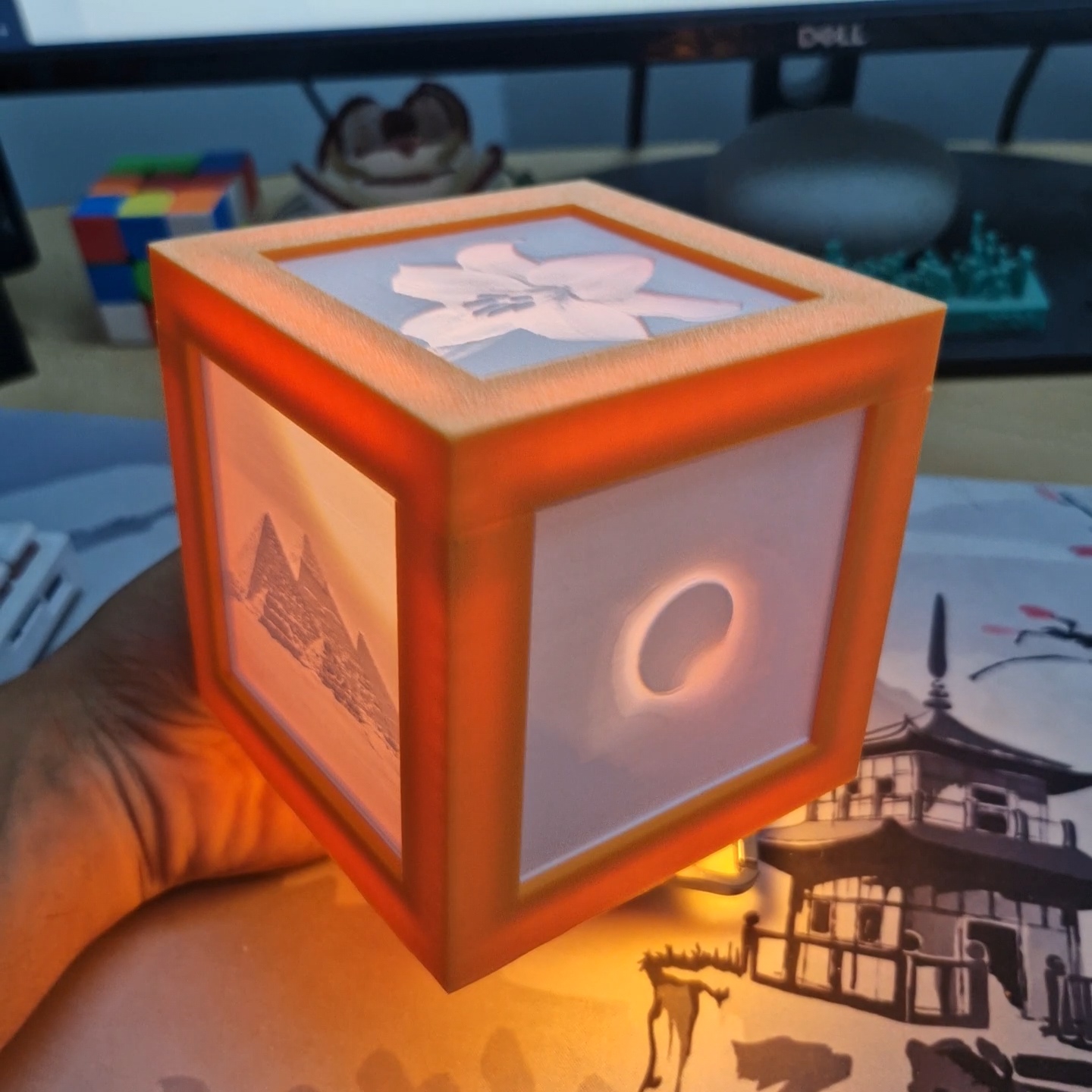 A small cube lamp with an orange frame, lit from inside. A white lily glows on the top panel, the pyramids of Giza on the left side and a solar eclipse on the right.